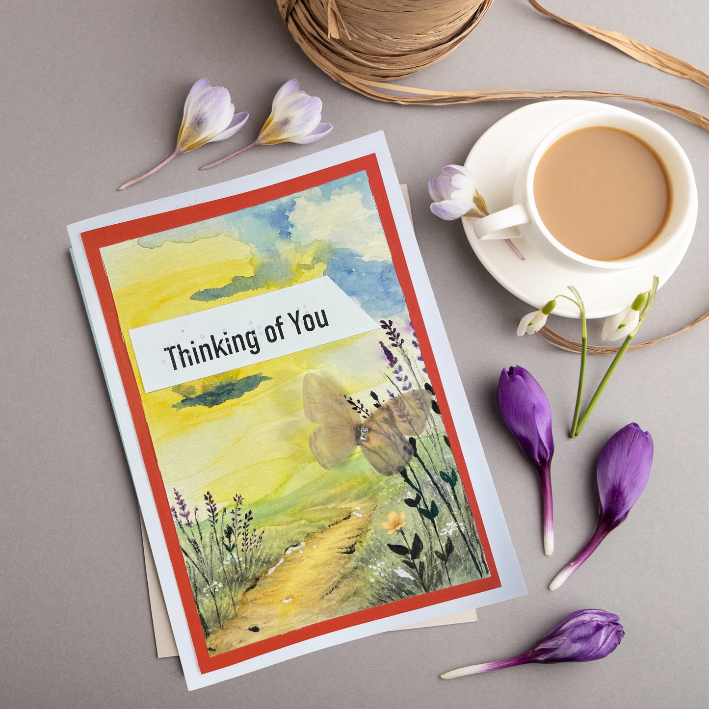 A greeting card with a watercolor landscape design and the text 'Thinking of You' is displayed in a styled flat-lay arrangement alongside a cup of coffee, flowers, and twine on a neutral gray background.