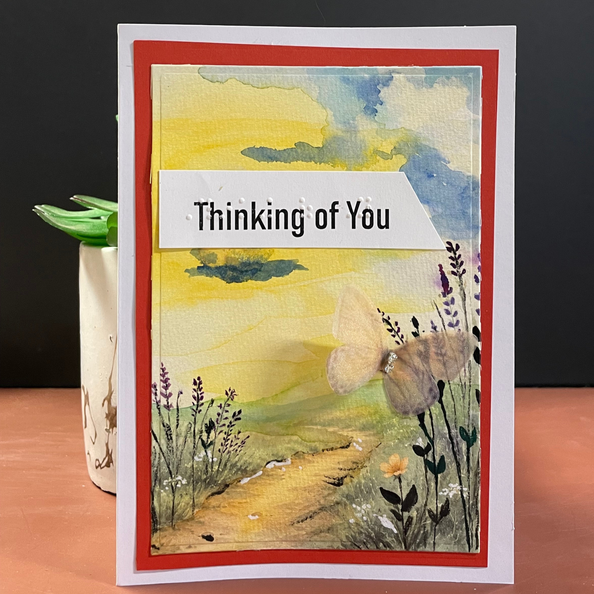 A handmade greeting card with a watercolor landscape design, featuring a yellow and blue sky, a butterfly, wildflowers, and a path. The card has a red border, a white inner border, and a white banner across the middle with the text 'Thinking of You'.