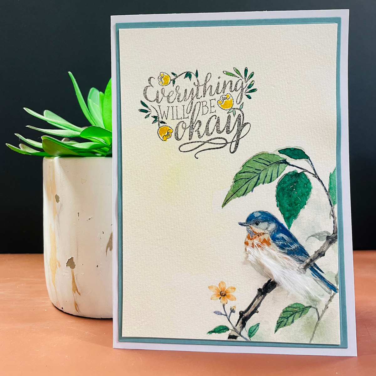 A handmade greeting card with a watercolor bluebird perched on a branch and the phrase 'Everything will be okay' in script, styled on a wooden surface next to a potted succulent against a dark background.