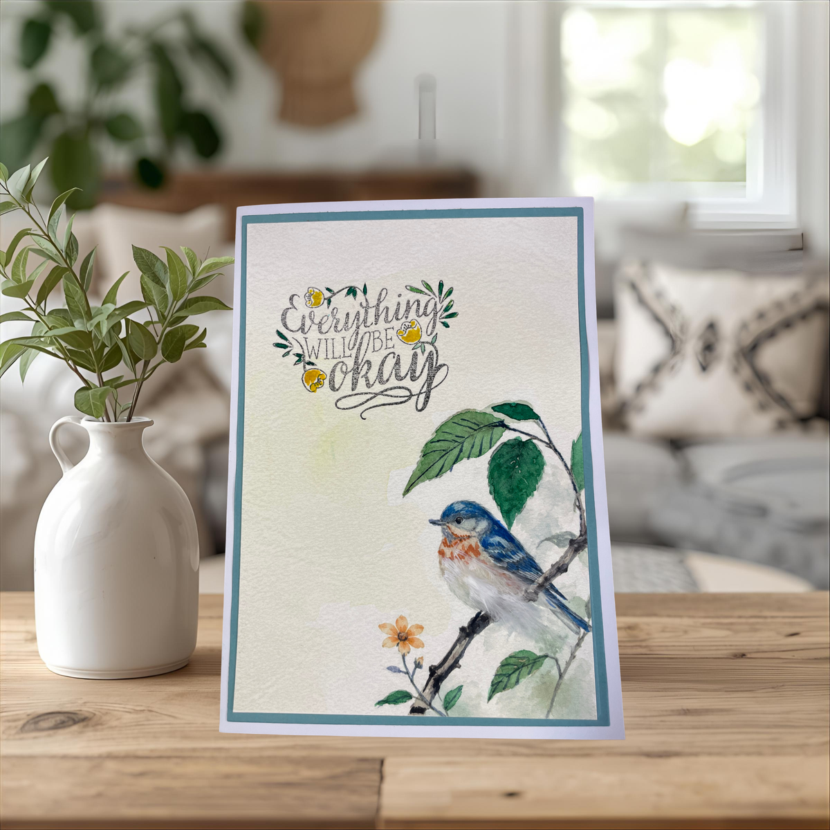 A handmade greeting card featuring a watercolor bluebird perched on a branch with the phrase 'Everything will be okay' in script, styled on a wooden table next to a white ceramic vase with green leaves.