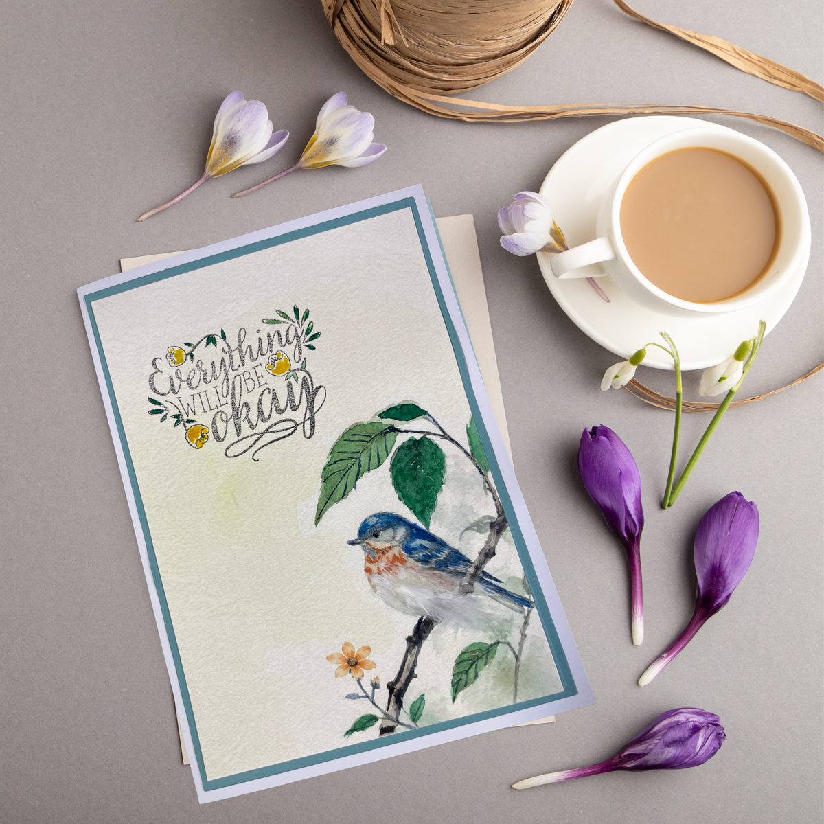 A greeting card with a watercolor robin and the phrase 'Everything is okay' in script, styled in a flat lay with a cup of coffee, purple flowers, and twine on a gray surface.