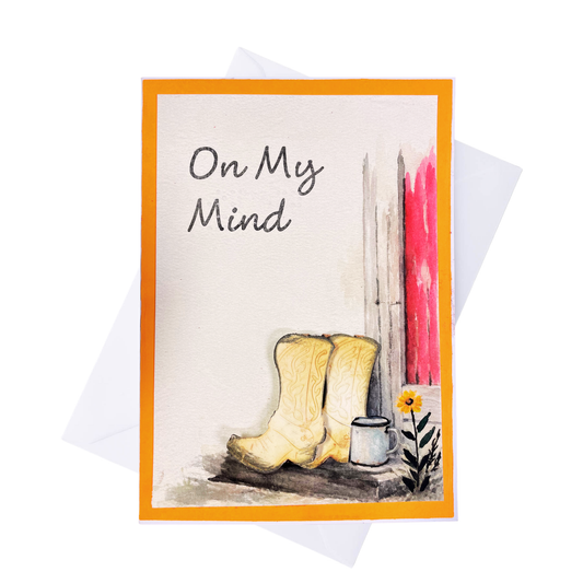 A greeting card with an orange border and the handwritten text and braille 'On My Mind,' featuring a watercolor-style illustration of off white cowboy boots, a white mug, a small yellow flower, and a red vertical brushstroke on a white background. A white envelope is partially visible behind the card.