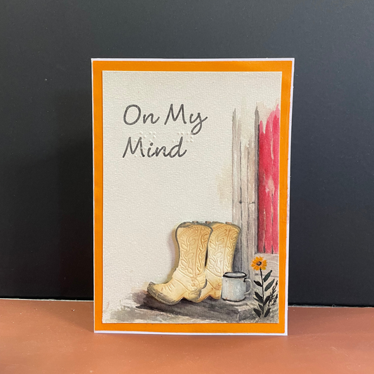 A handmade greeting card with an orange border, featuring the text and braille  'On My Mind' above a watercolor of beige cowboy boots, a small white cup, and a yellow flower against a off white background.
