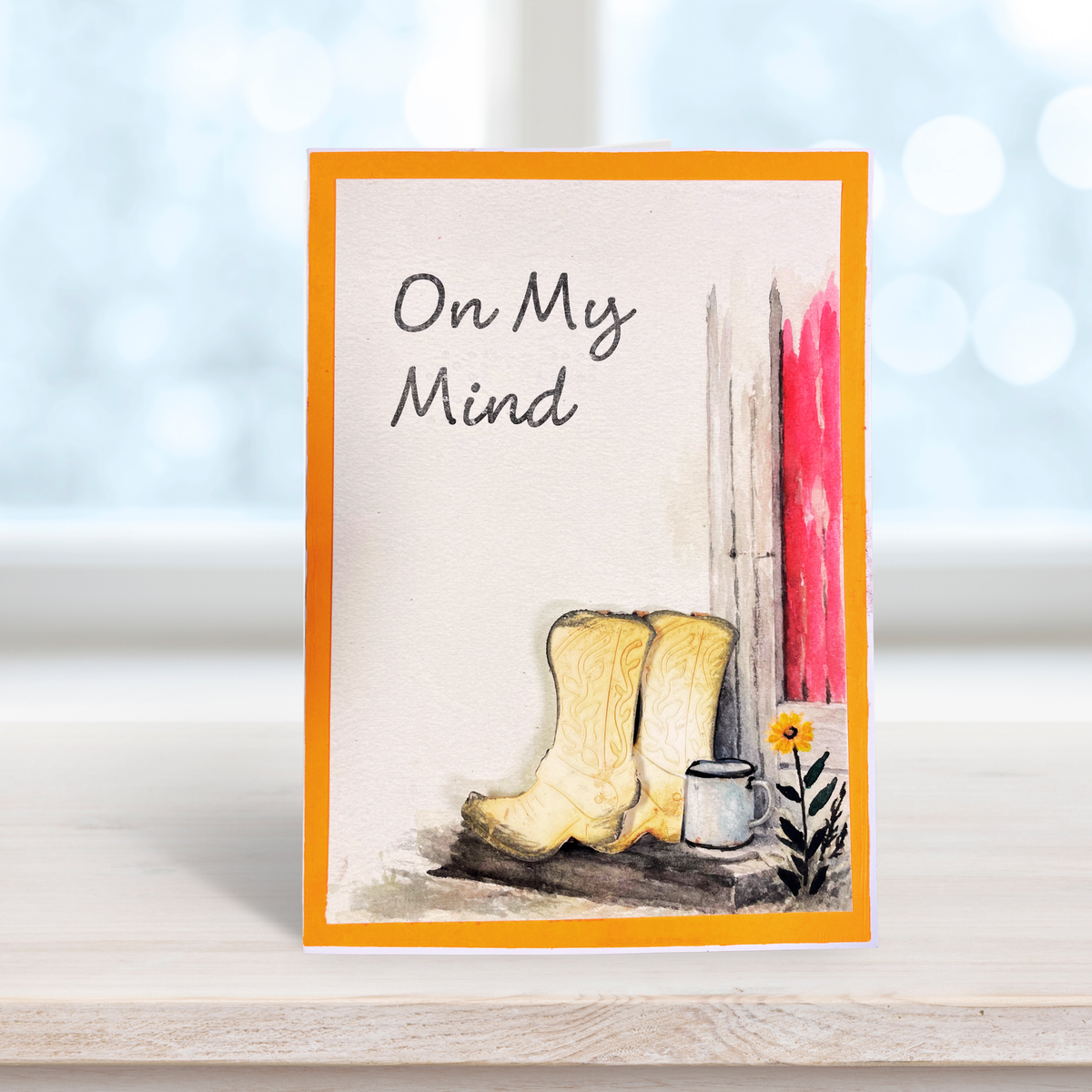 A handmade greeting card with an orange border, featuring the text and braille  'On My Mind' above a watercolor of beige cowboy boots, a small white cup, and a yellow flower against a off white background.