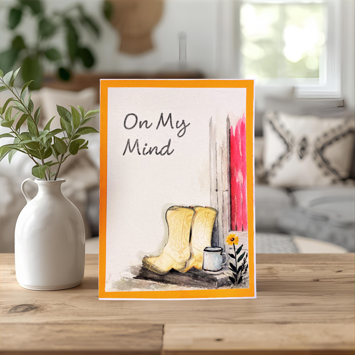 A handmade greeting card with an orange border, featuring the text and braille  'On My Mind' above a watercolor of beige cowboy boots, a small white cup, and a yellow flower against a living room background.