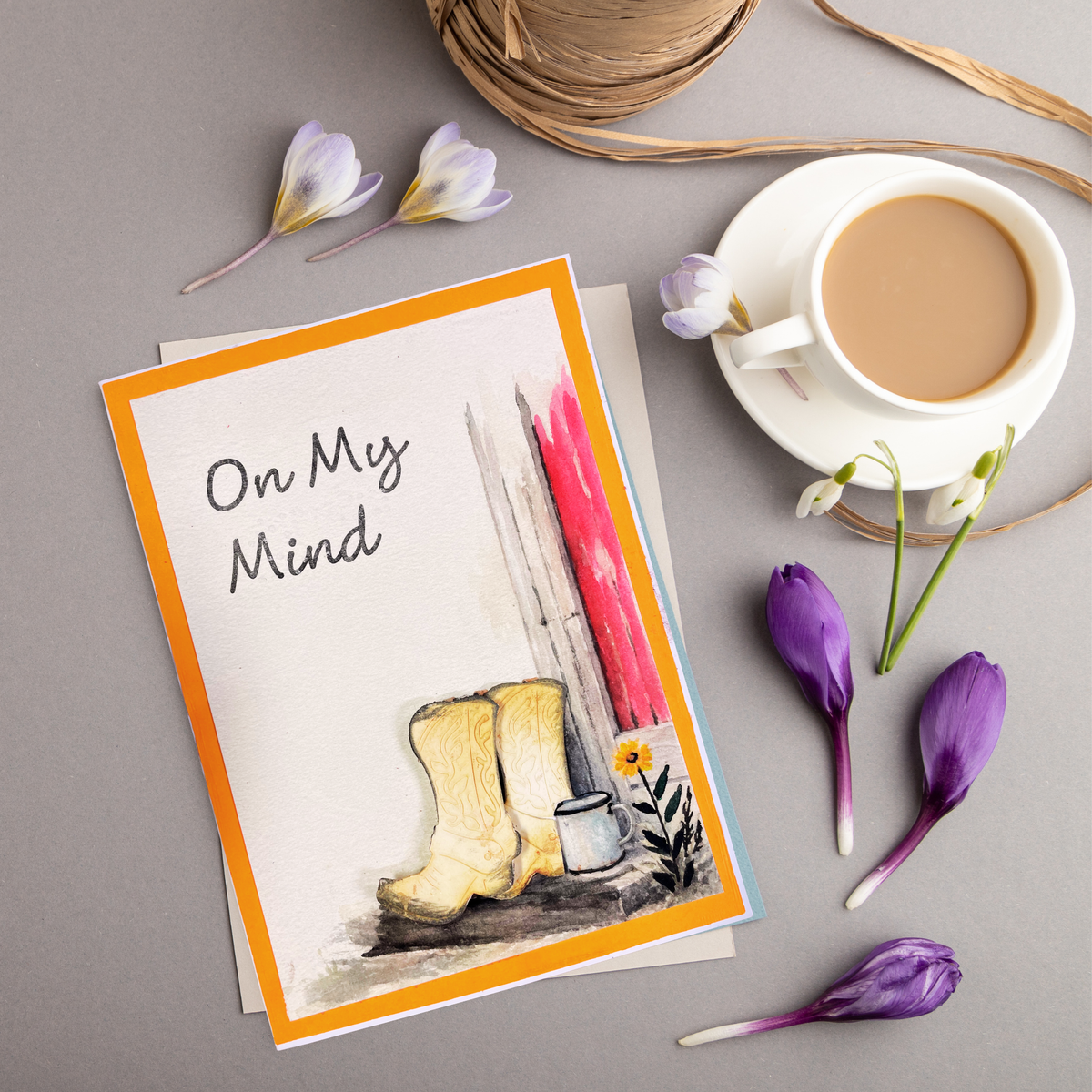 A handmade greeting card with an orange border, featuring the text and braille  'On My Mind' above a watercolor of beige cowboy boots, a small white cup, and a yellow flower against  background styled on a gray surface with a cup of coffee, twine, and purple flower petals.