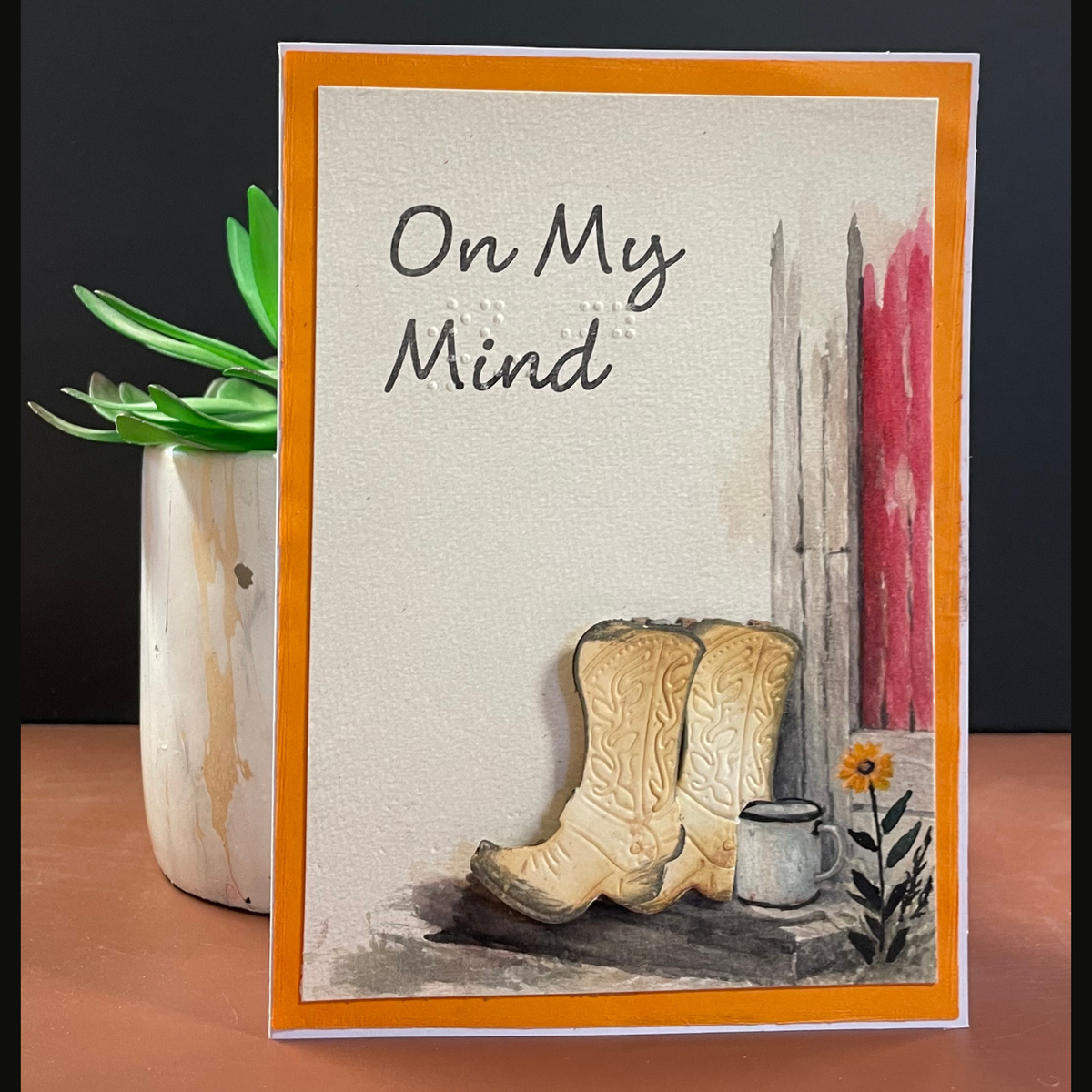 A handmade greeting card with an orange border, featuring the text and braille  'On My Mind' above a watercolor of beige cowboy boots, a small white cup, and a yellow flower against a off white background.