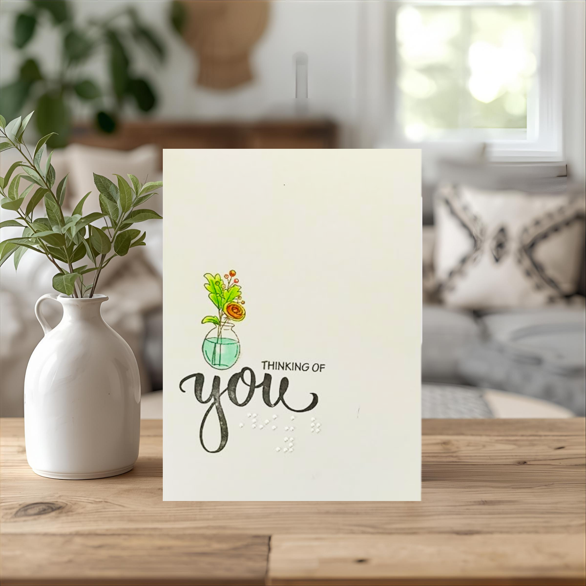 A greeting card with a watercolor illustration of flowers in a glass vase and the words 'THINKING OF you' in black script and braille displayed on a wooden table next to a white ceramic vase with green leaves.