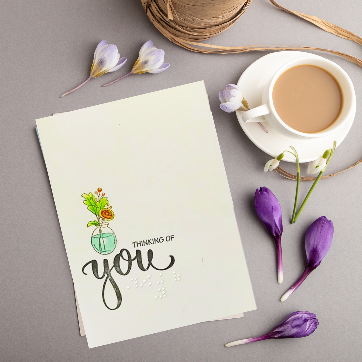 A cream-colored greeting card with a watercolor illustration of flowers in a vase and the text 'THINKING OF you' in black script and braille, styled in a flat-lay with a cup of coffee, purple flowers, and twine on a gray background.