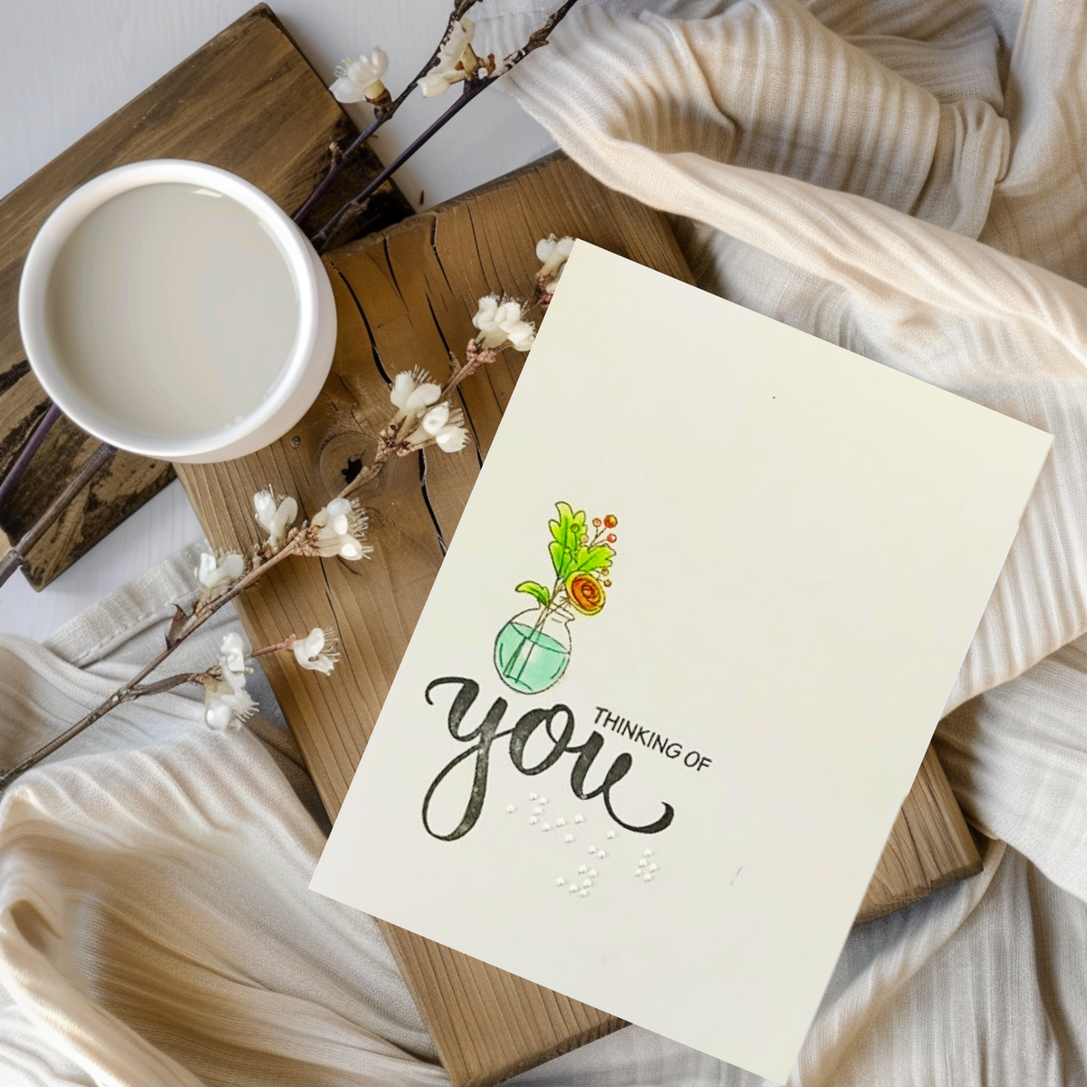 A greeting card with a colorful illustration of a flower in a vase and the text 'THINKING OF you' in black script and braille, styled in a flat-lay arrangement on a wooden board with a cup of coffee, dried flowers, and beige ribbed fabric.
