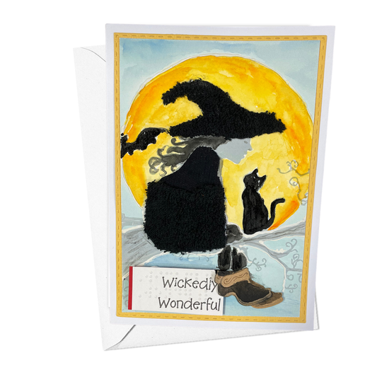 A Halloween greeting card with a white envelope behind it, featuring a stylized illustration of a witch in a black hat and cloak with a black cat in front of a large yellow moon, and the words 'Wickedly Wonderful' on the card.