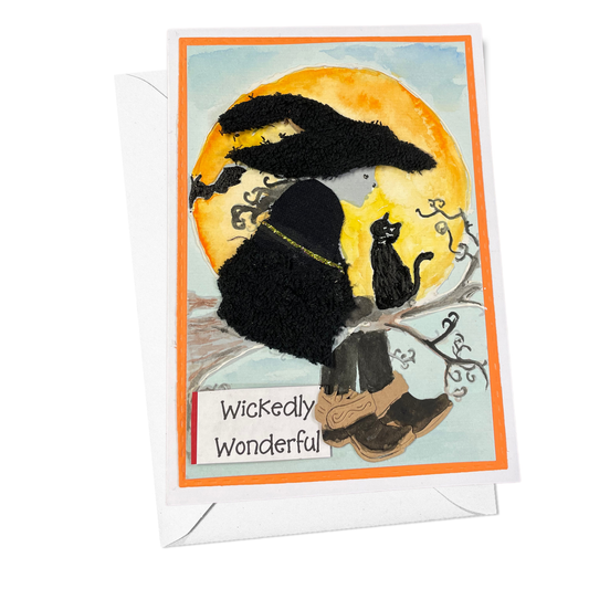 A greeting card with a white envelope behind it, featuring a whimsical Halloween illustration of a witch in black clothing sitting on a branch in front of a large yellow moon, with a black cat and the words 'Wickedly Wonderful' on the card.